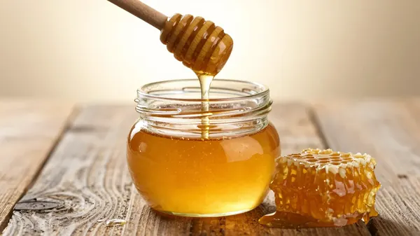 Golden honey dripping from a wooden dipper into a glass jar beside honeycomb